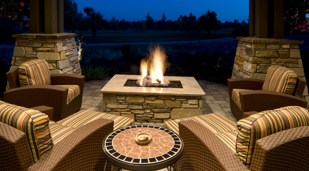 square stone fire pit lit up at nigh surrounded by patio chairs under cover